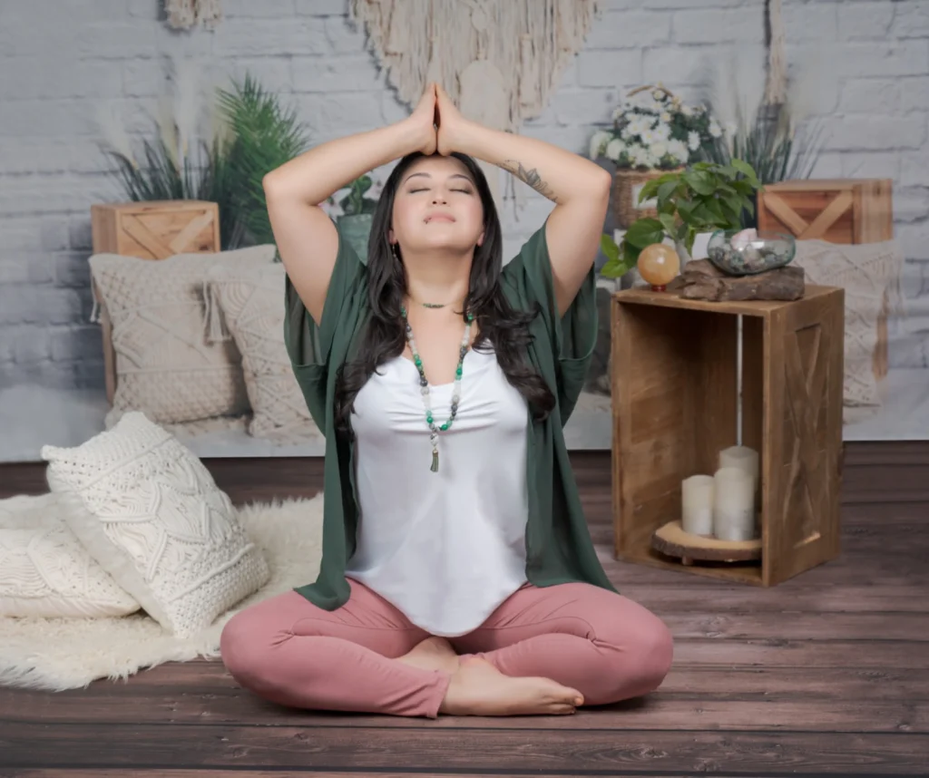 Woman sitting cross-legged in a calm studio space, eyes closed with hands pressed together above her head in a yoga or meditation pose, surrounded by soft pillows, plants, candles, and natural décor.